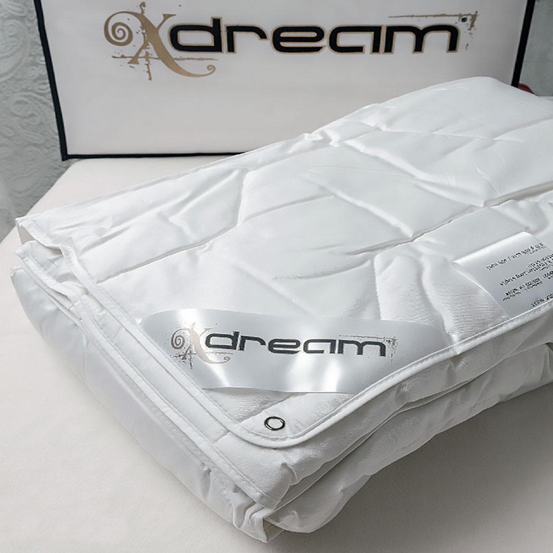 Белое одеяло X-DREAM сложено стопкой, видна атласная бирка с логотипом и пришитая бирка состава, позади фирменная упаковка X-DREAM