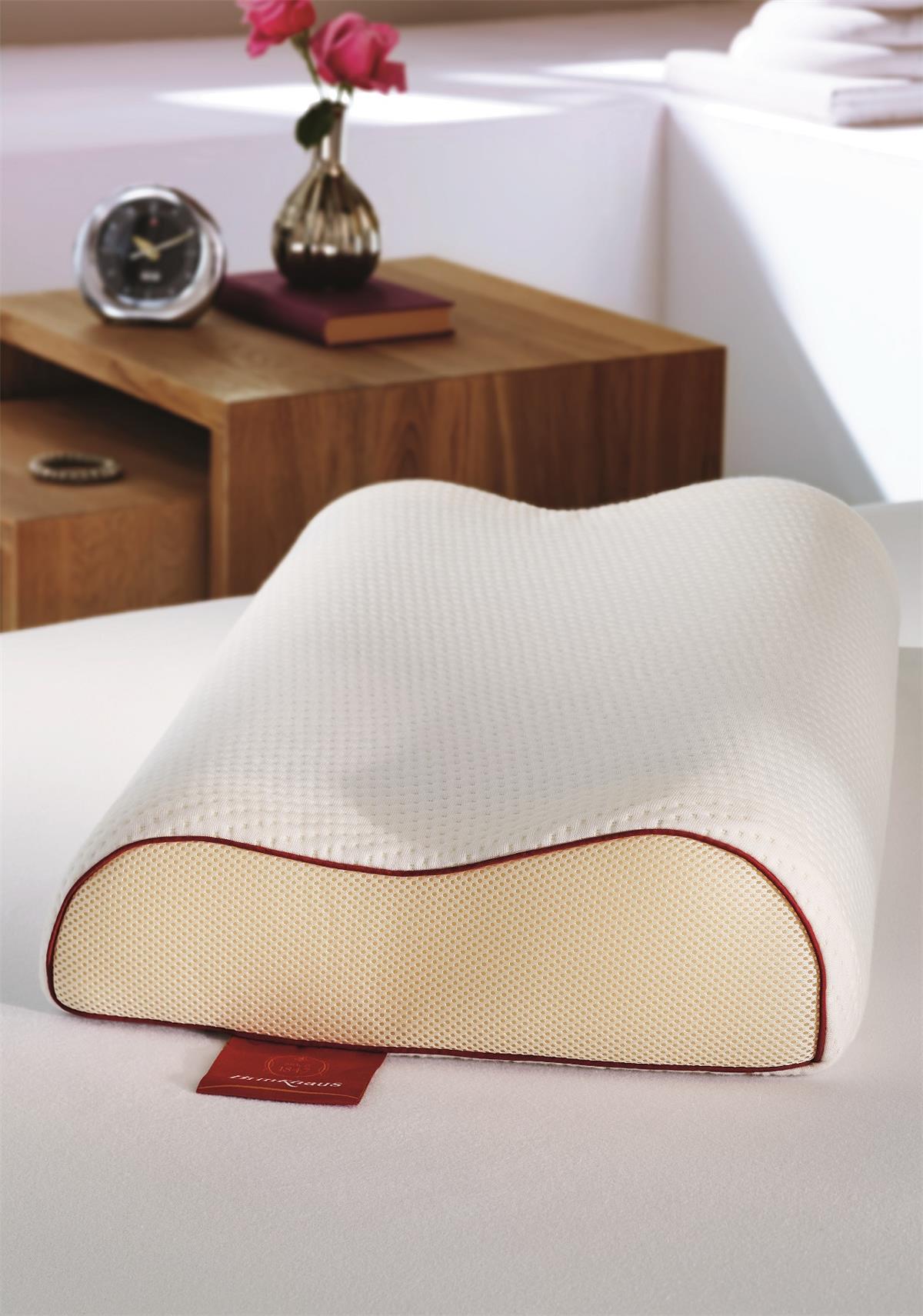 Подушка Brinkhaus THE GEL NECK SUPPORT на белом матрасе: анатомическая волна, верх из белого объёмного трикотажа, боковая вставка кремовой сетки, бордовый кант по краю, красная фирменная бирка Brinkhaus снизу; за подушкой деревянный ночник с настольными часами, книгой и розами в латунной вазе