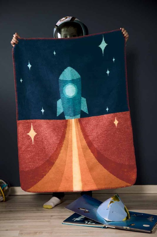 Ребёнок держит перед собой детский плед Biederlack LOVELY SWEET LITTLE ROCKET с ракетой, на полу игрушечный шлем космонавта