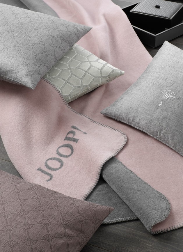 Плед JOOP! ROSE GRAPHIT, розово-серый, с вышитым логотипом JOOP!, разложен на кровати рядом с декоративными подушками той же коллекции