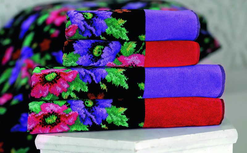 Четыре шенилловых полотенца Feiler MOHN SCHWARZ сложены стопкой на белой тумбе: чёрное поле с розовыми маками и синими васильками, бордюры фиолетовый и красный