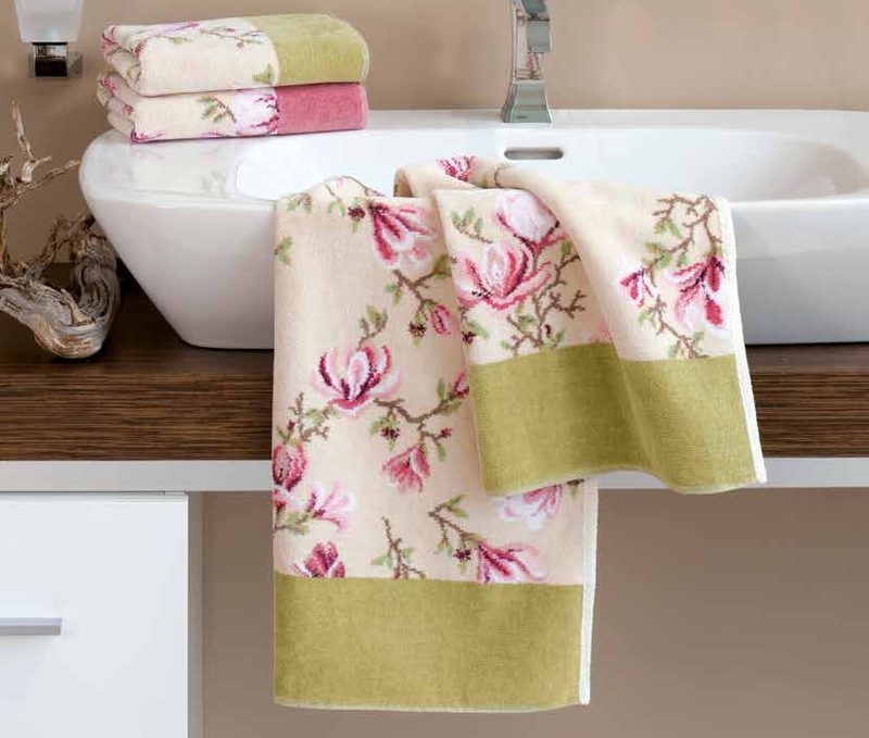 Шенилловое полотенце Feiler MAGNOLIA BEIGE с розовыми магнолиями на кремовом поле и оливковым бордюром перекинуто через деревянную столешницу у белой раковины, слева сложены полотенца того же рисунка